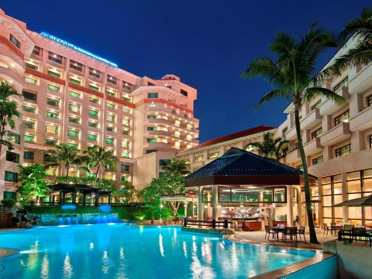 Paradox Singapore Merchant Court At Clarke Quay Экстерьер фото The photo depicts a luxurious hotel complex set against a twilight sky. In the foreground, there is a large swimming pool that reflects the light from surrounding lights and the sky. Lush greenery, including palm trees, adds to the tropical ambiance.