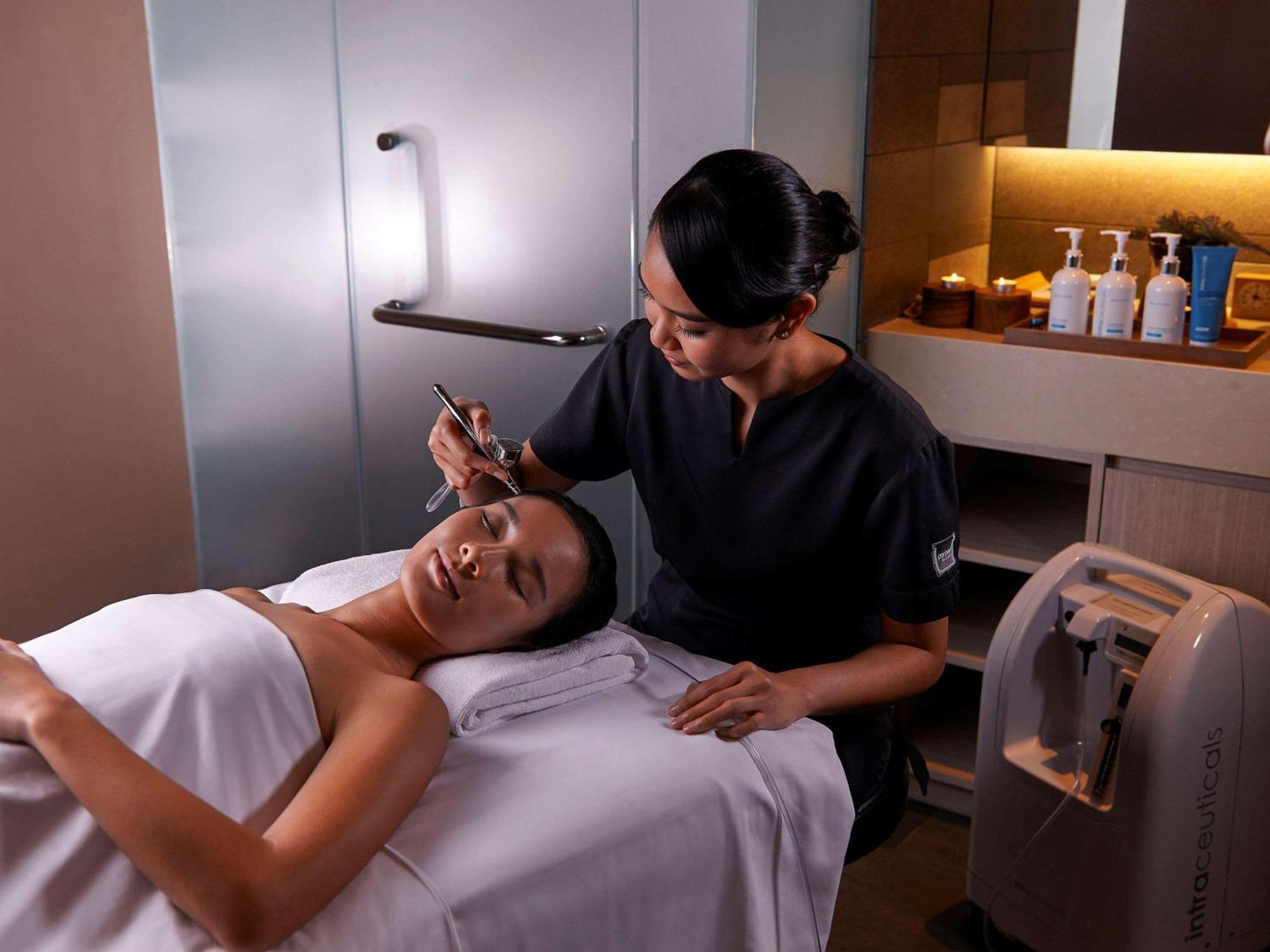 Paradox Singapore Merchant Court At Clarke Quay Экстерьер фото The photo shows a spa or beauty therapy setting where a woman is lying on a treatment table with a towel draped over her. A professional, dressed in a dark uniform, is performing a skincare treatment on her face. Various beauty products are visible o