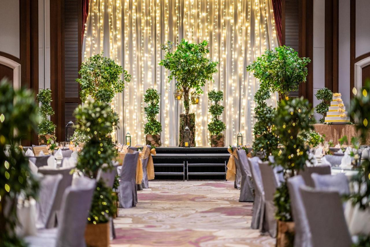 Paradox Singapore Merchant Court At Clarke Quay Экстерьер фото The image shows an elegantly decorated event space, likely for a wedding or formal celebration. In the center, there is a backdrop with draped fabric illuminated by soft lights, creating a warm ambiance. There are several decorative trees or potted p