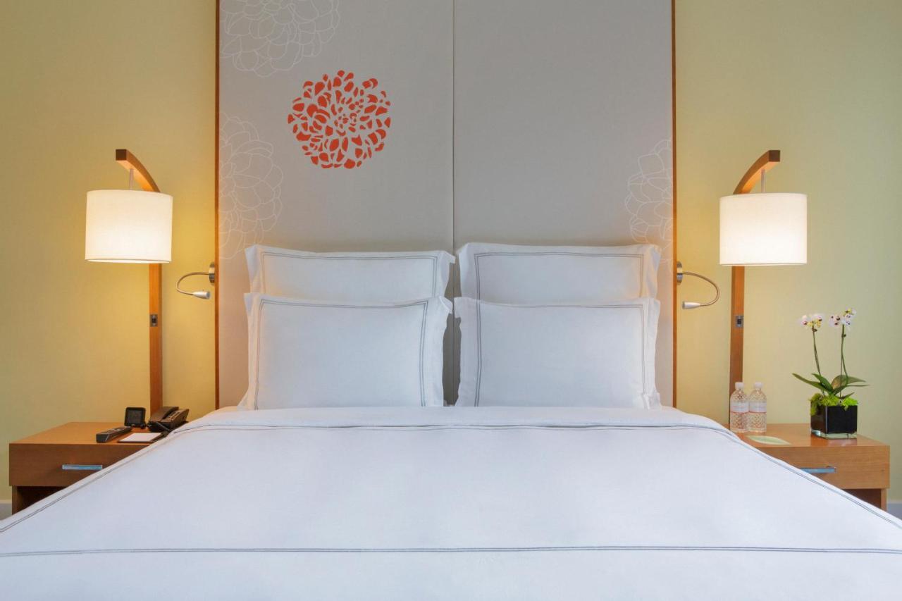 Paradox Singapore Merchant Court At Clarke Quay Экстерьер фото The photo showcases a cozy hotel room with a neatly made bed featuring white linens and two pillows. Behind the bed is a decorative headboard with a simple floral design in orange. On either side of the bed, there are stylish bedside lamps providing 