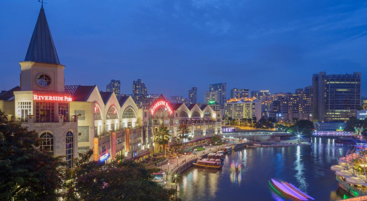 Paradox Singapore Merchant Court At Clarke Quay Экстерьер фото The photo shows a riverside scene at dusk, featuring a waterfront area with modern buildings that have a distinctive architectural style. The structures have pointed roofs and are well-lit, creating a vibrant atmosphere. Along the river, there are bo