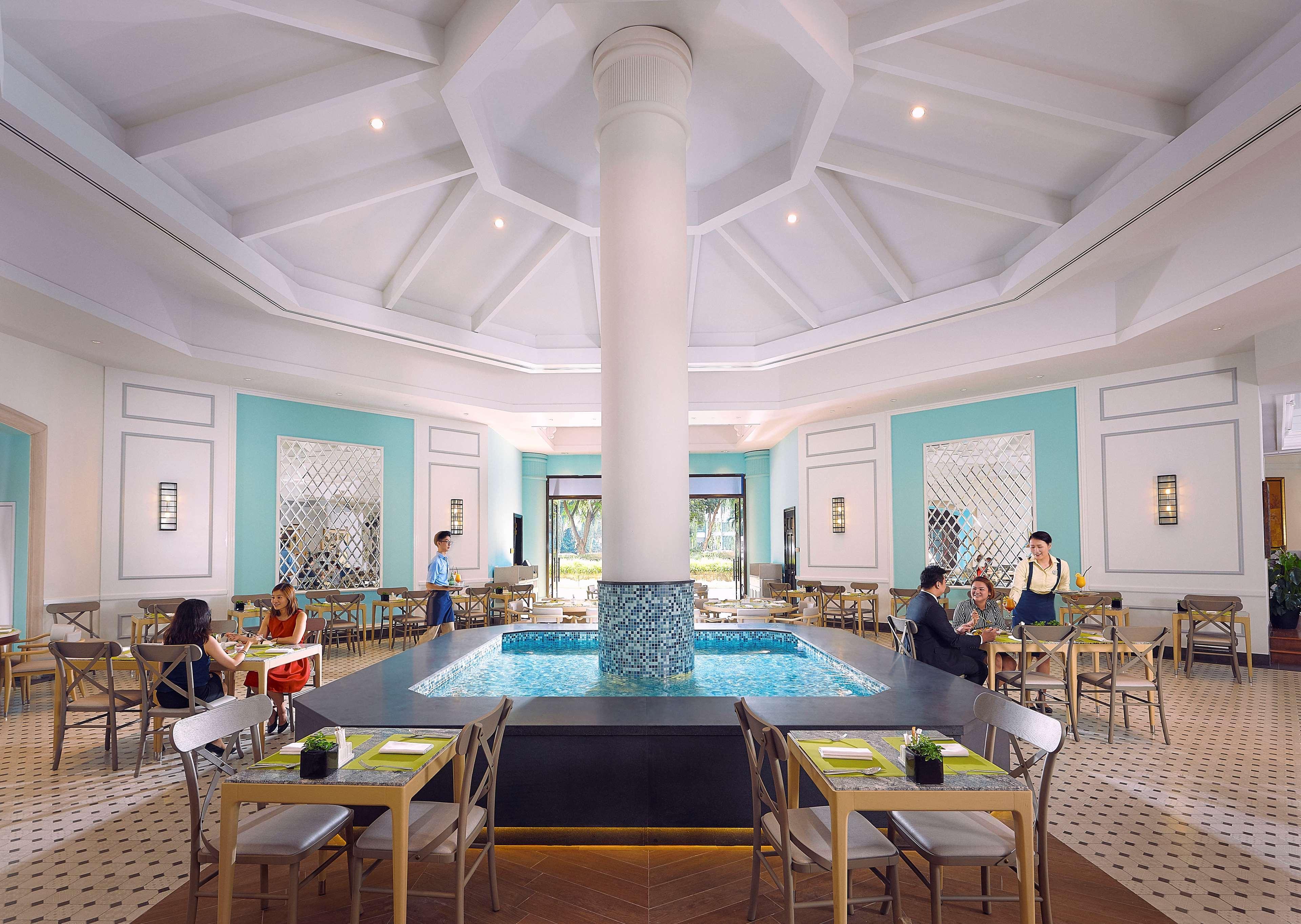 Paradox Singapore Merchant Court At Clarke Quay Экстерьер фото The photo depicts an elegant restaurant interior characterized by a spacious, well-lit design. At the center, there is a large, round column that supports a high, intricately designed ceiling. Surrounding the column is a small pool of water, adding a