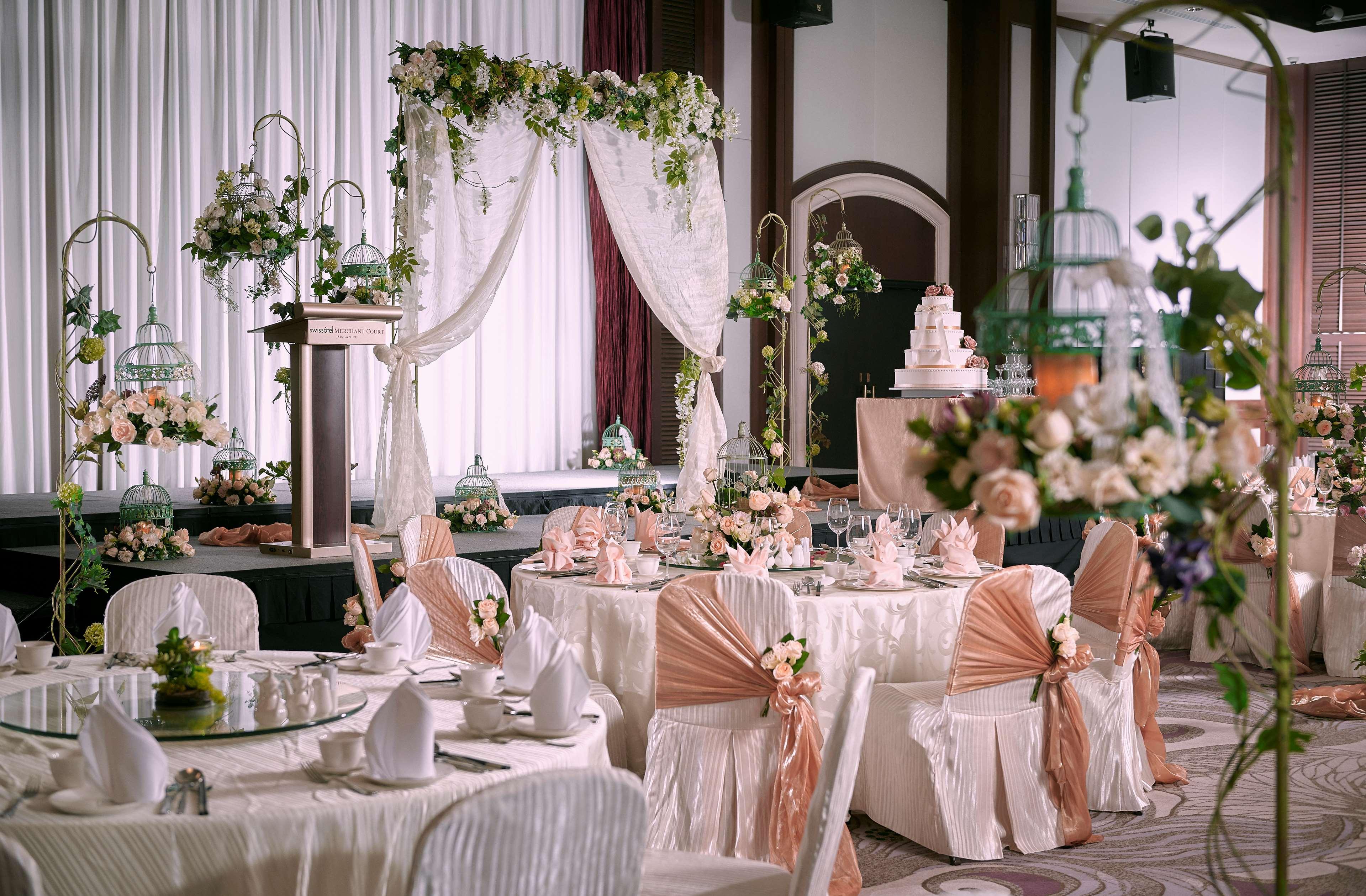 Paradox Singapore Merchant Court At Clarke Quay Экстерьер фото The photo shows an elegantly decorated indoor event space, likely set up for a wedding or a formal celebration. There are several round tables covered with white tablecloths, each adorned with floral arrangements and cloth napkins. The chairs are dre