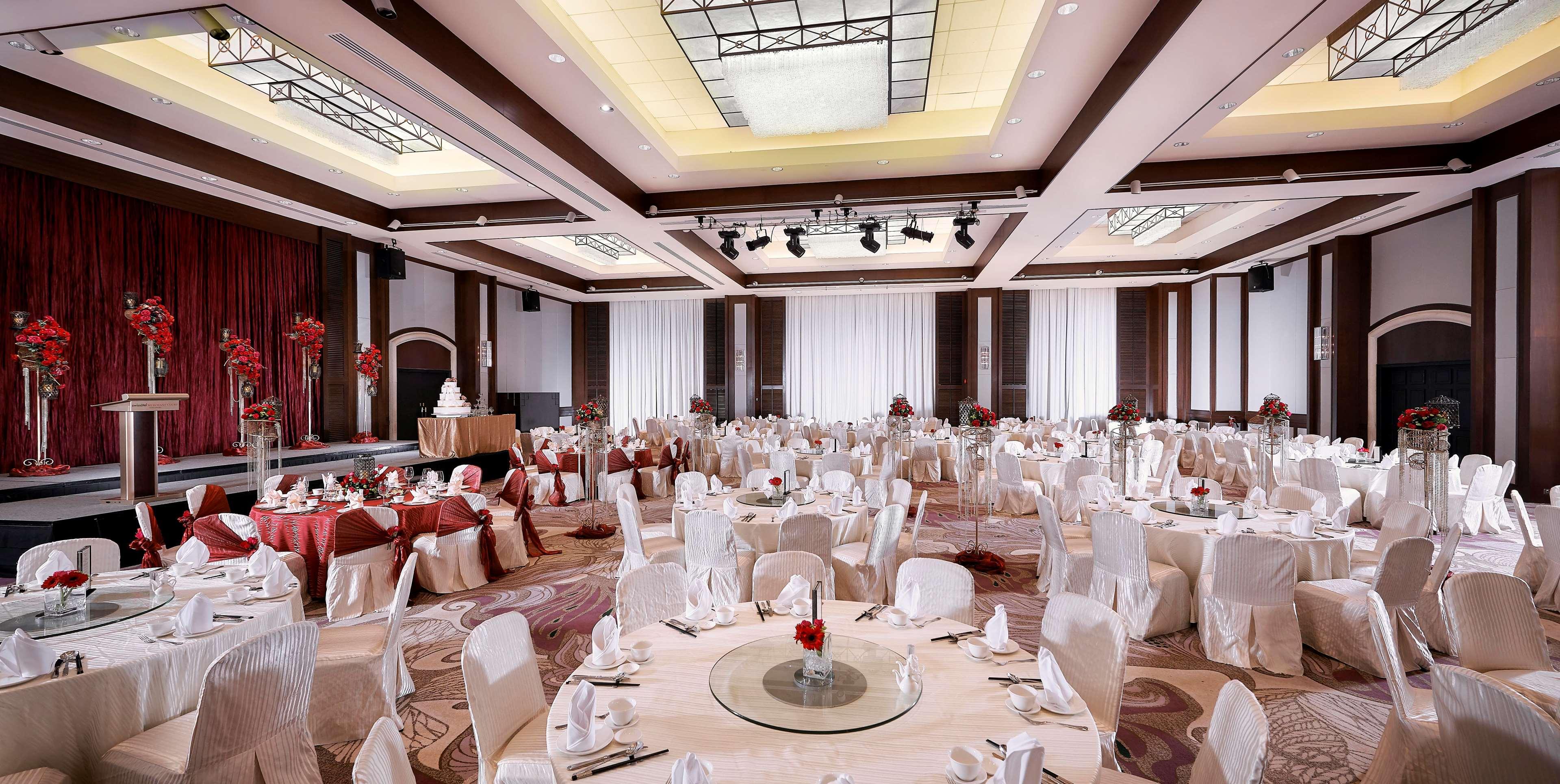 Paradox Singapore Merchant Court At Clarke Quay Экстерьер фото The photo shows a spacious banquet hall set up for an elegant event. Round tables are beautifully arranged, each covered with a tablecloth and adorned with a center decoration featuring red flowers. The chairs are draped in white covers, adding to th