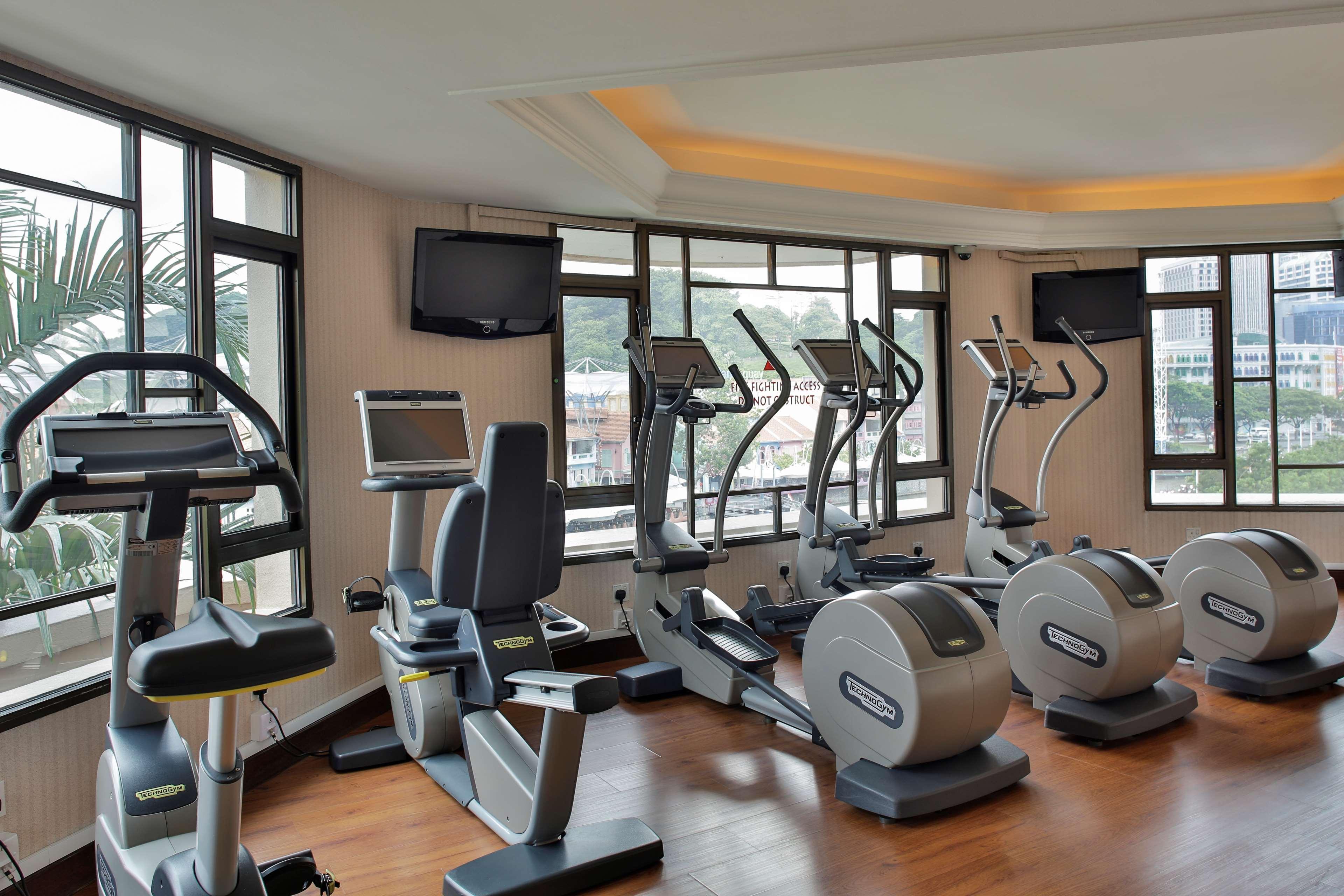 Paradox Singapore Merchant Court At Clarke Quay Экстерьер фото The photo shows a fitness room equipped with various exercise machines. There are several elliptical machines and stationary bicycles positioned by large windows, allowing for natural light to fill the space. Additionally, there are two televisions m