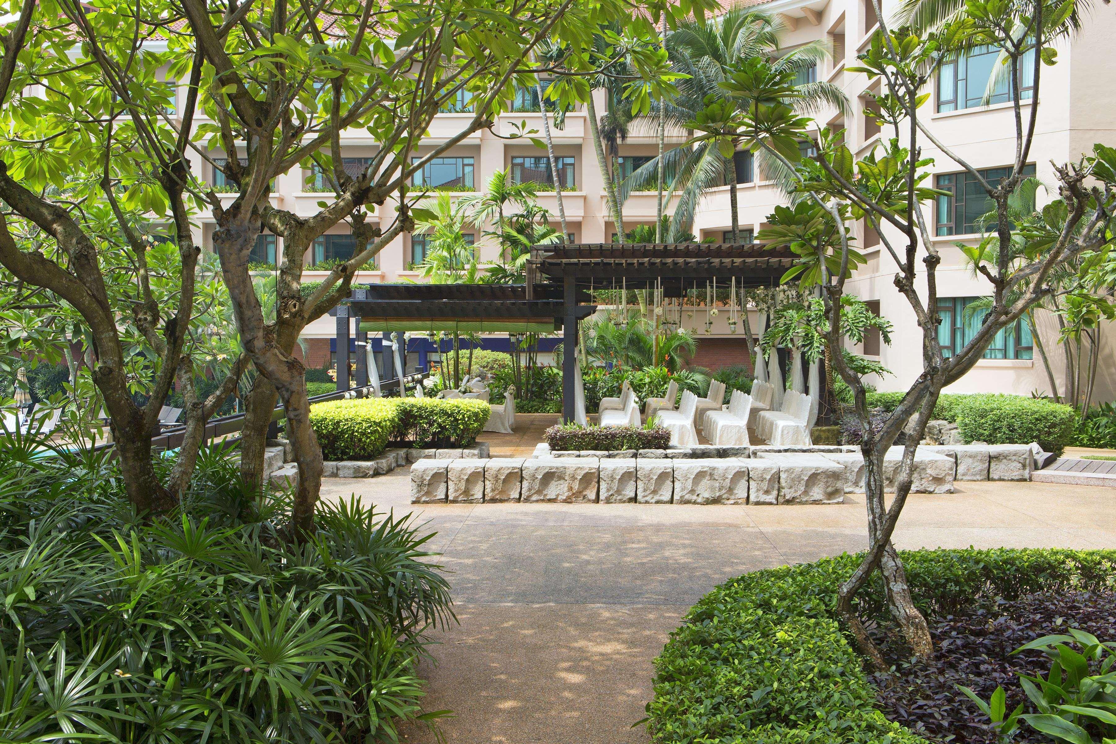 Paradox Singapore Merchant Court At Clarke Quay Экстерьер фото The photo depicts a garden or courtyard area surrounded by lush greenery. There are several trees and vibrant plants, creating a serene environment. In the center, there seems to be a stone seating arrangement under a covered structure, likely meant 