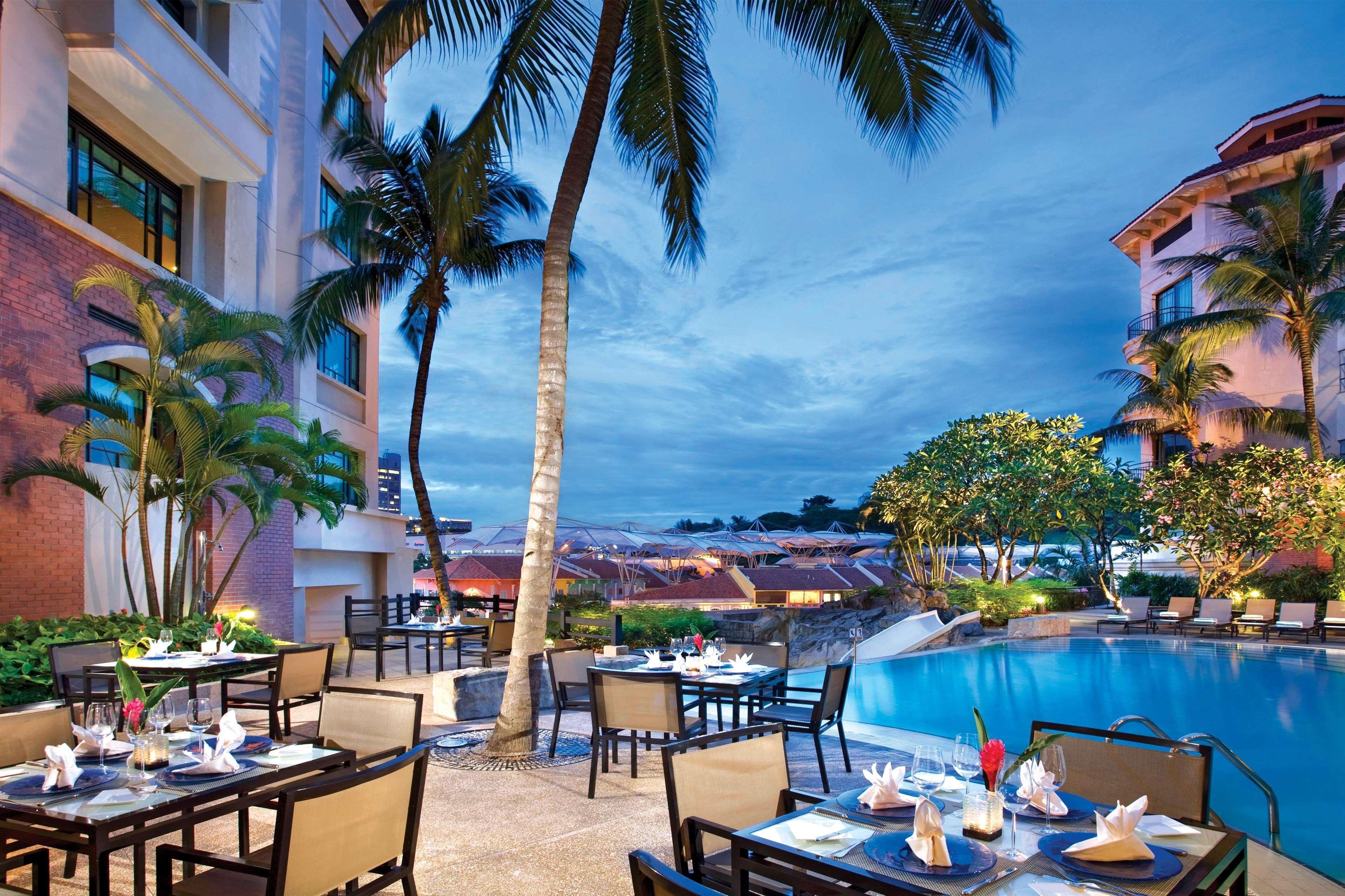 Paradox Singapore Merchant Court At Clarke Quay Ресторан фото The photo depicts a beautiful outdoor dining area next to a shimmering pool, set against a backdrop of palm trees and a dramatic evening sky. Tables are arranged elegantly with white tablecloths, chairs, and decorative napkins, suggesting a luxurious