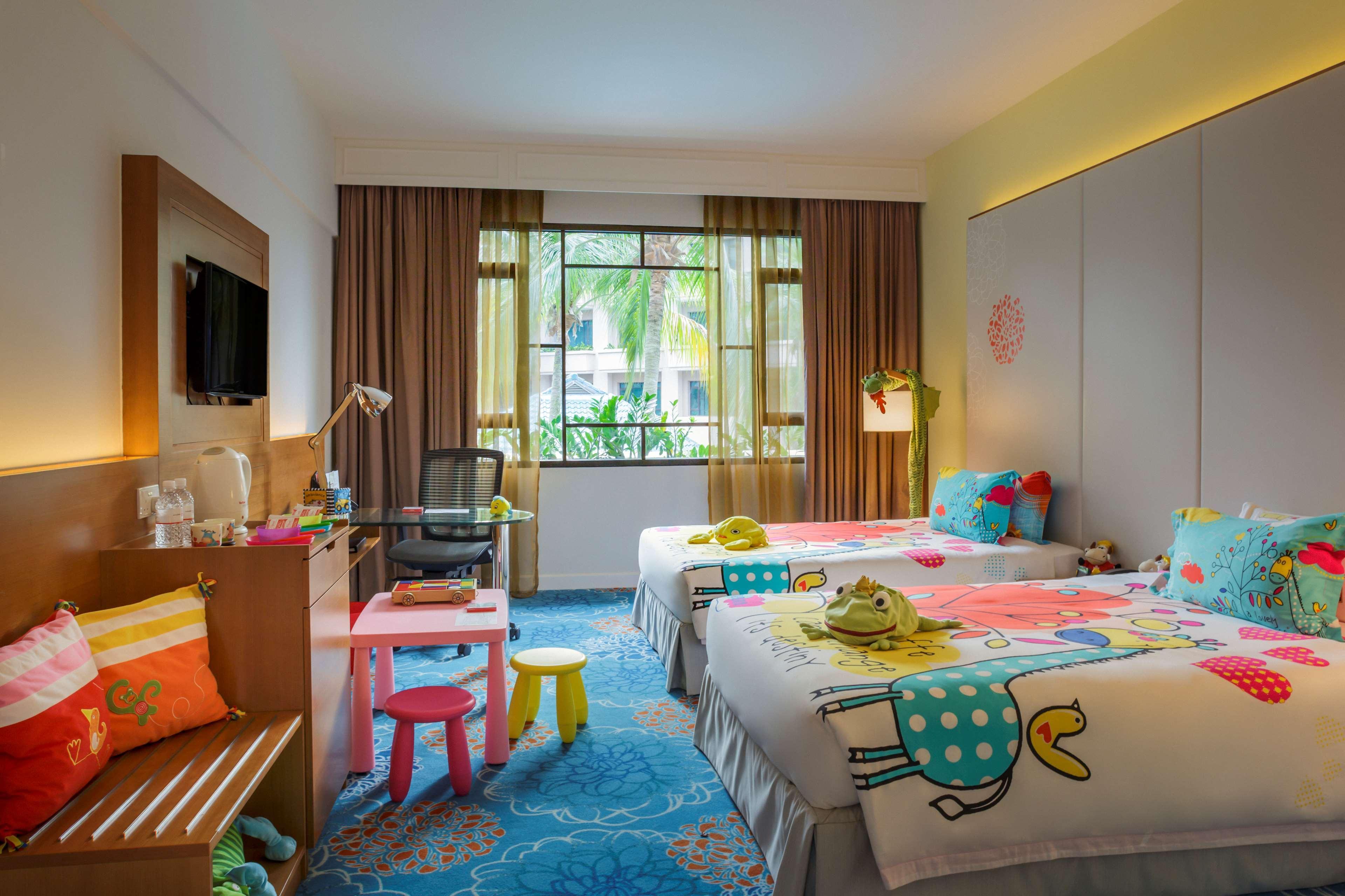 Paradox Singapore Merchant Court At Clarke Quay Экстерьер фото The photo shows a brightly colored hotel room designed in a family-friendly style. There are two twin beds, each adorned with playful, colorful bedding featuring whimsical patterns. A small table with brightly colored stools is positioned nearby. A c