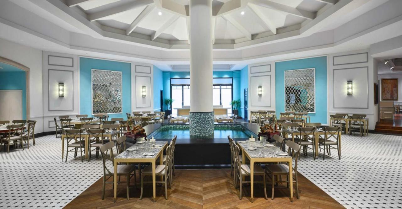 Paradox Singapore Merchant Court At Clarke Quay Экстерьер фото The photo depicts an elegantly designed restaurant interior. The space features a high, domed ceiling with a central pillar. The walls are painted in a vibrant blue, complemented by decorative mirrors and artwork. There are several tables arranged th
