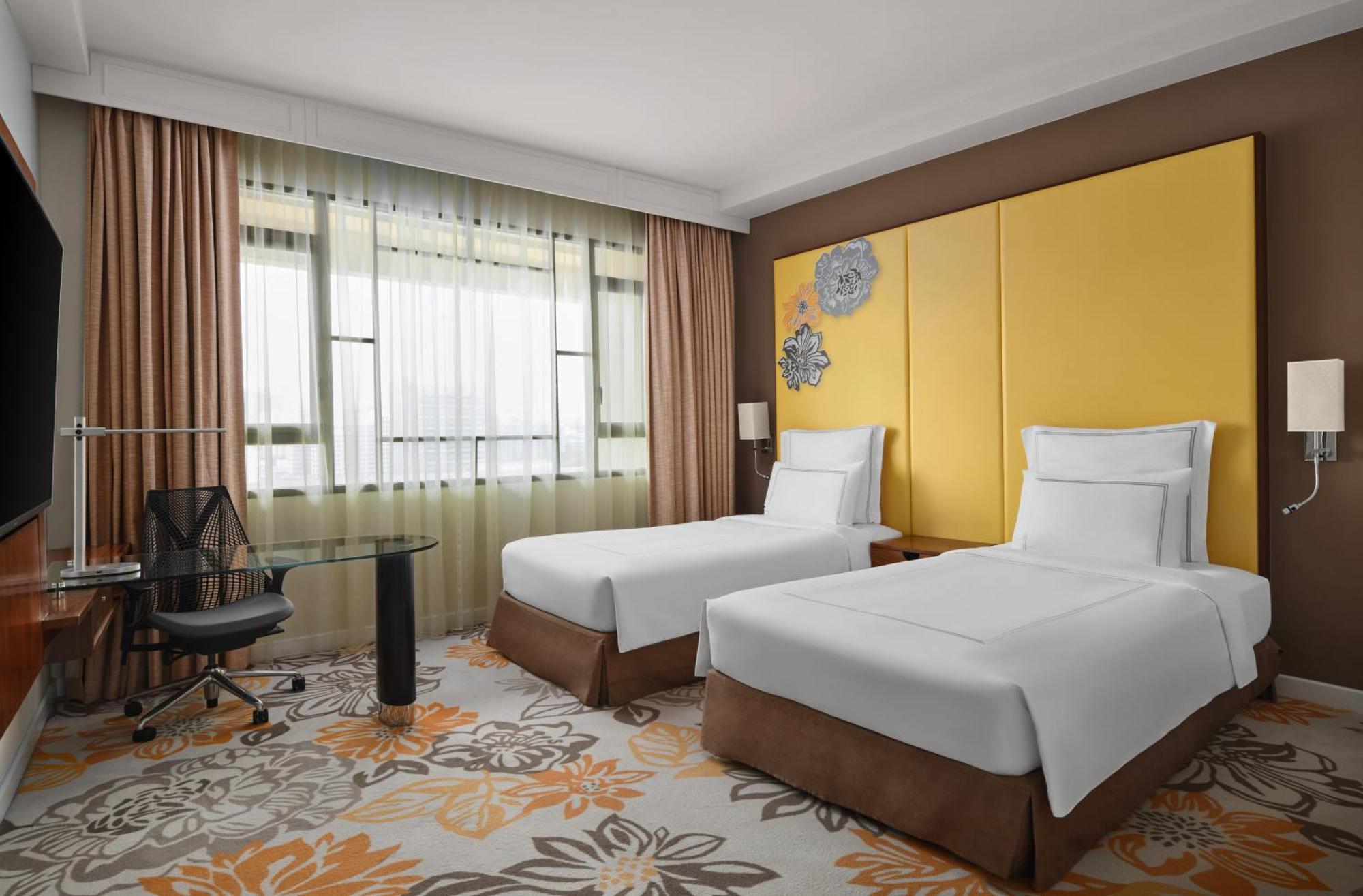 Paradox Singapore Merchant Court At Clarke Quay Экстерьер фото The photo shows a hotel room featuring two single beds with white linens and brown bed skirts. The room has a modern design with large windows dressed in sheer curtains, allowing natural light to enter. There is a small glass desk and an ergonomic ch